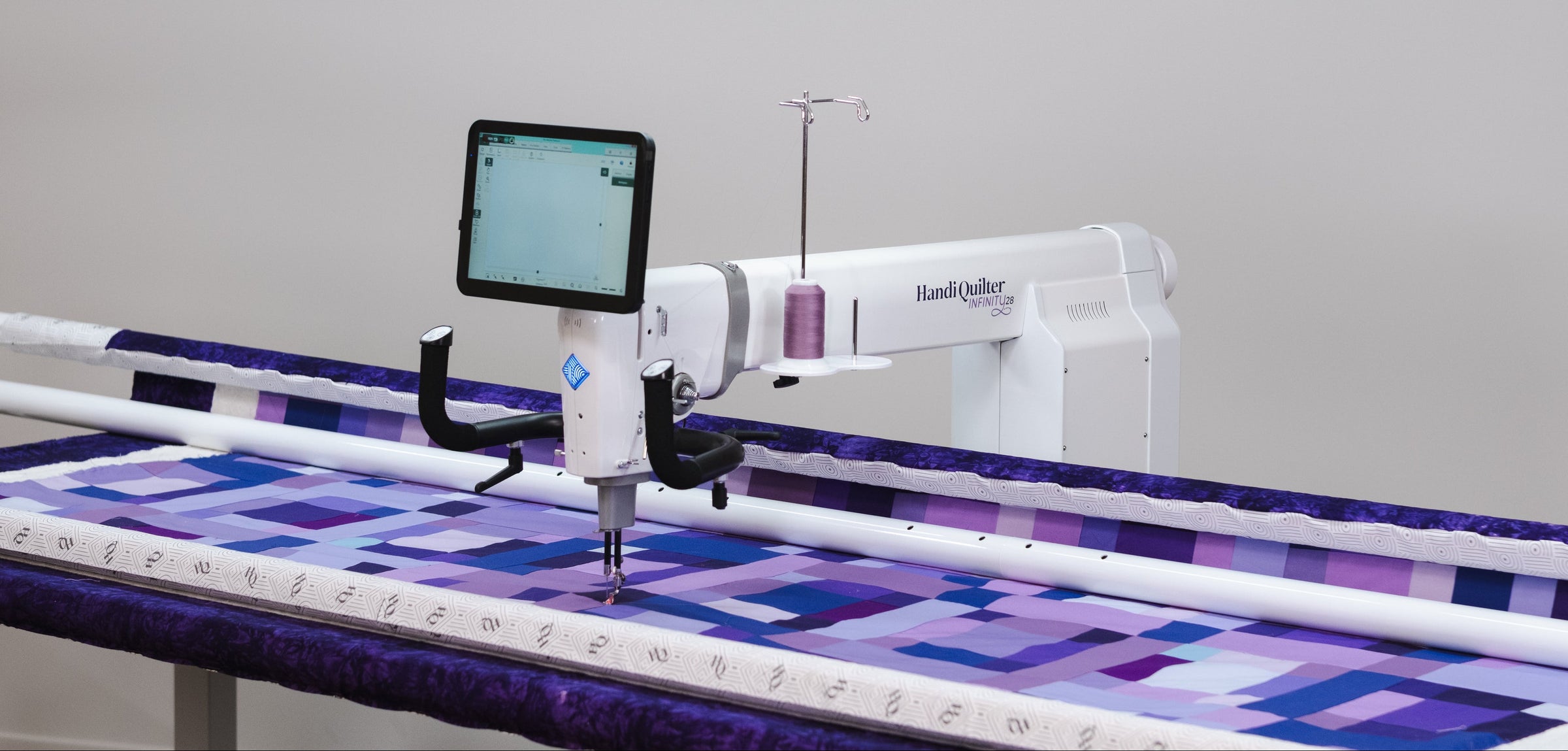 A white Handi Quilter longarm quilting machine with a digital touchscreen display, stitching a purple and white geometric patterned quilt on a large quilt frame under a bright overhead light bar.