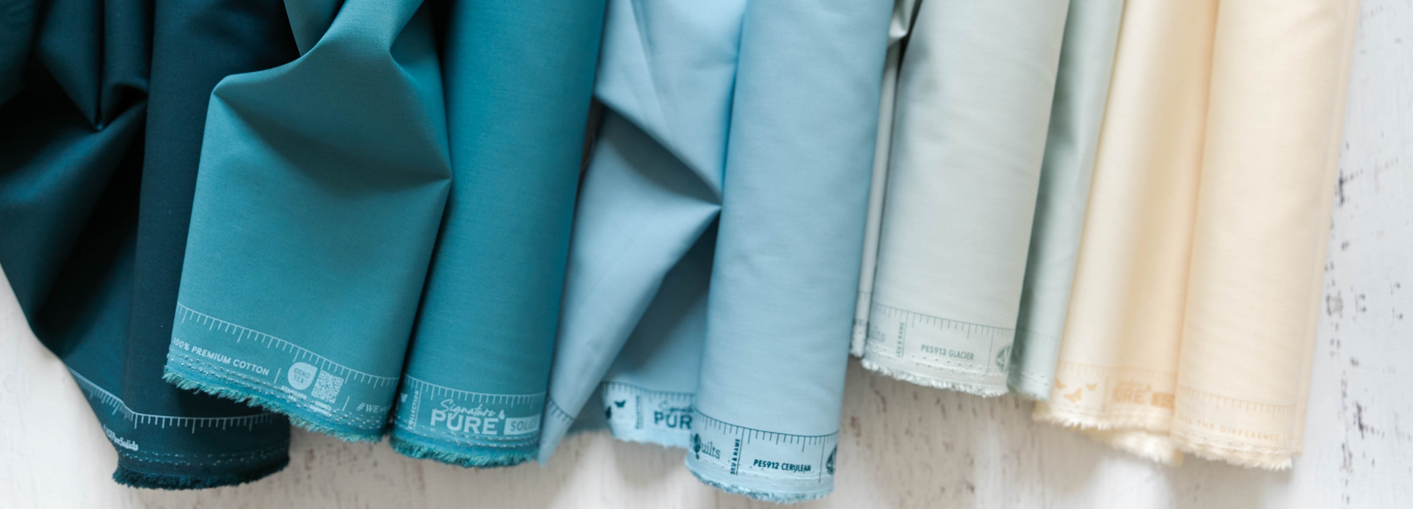 Several fabric rolls of Art Gallery Fabrics Signature Pure Solids laying side by side in muted teal, blue, pale gray, and cream tones against a white textured wall.