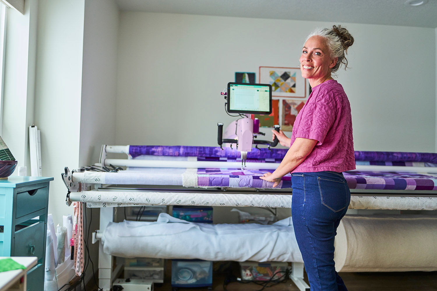 A woman in a pink shirt and blue jeans stands beside a large quilting machine with purple patterned fabric rolls in a bright craft room.