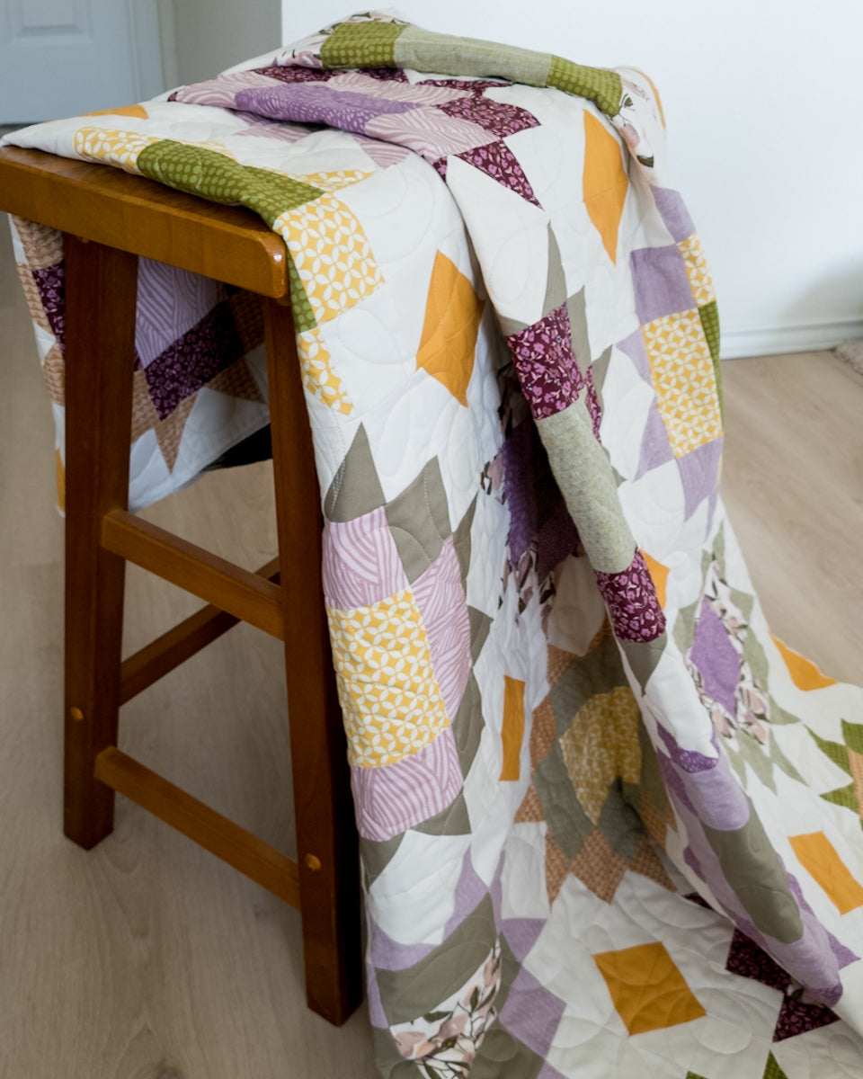 A patchwork quilt with geometric blocks in purple, olive green, mustard yellow, and gray draped over a wooden stool in a home setting.