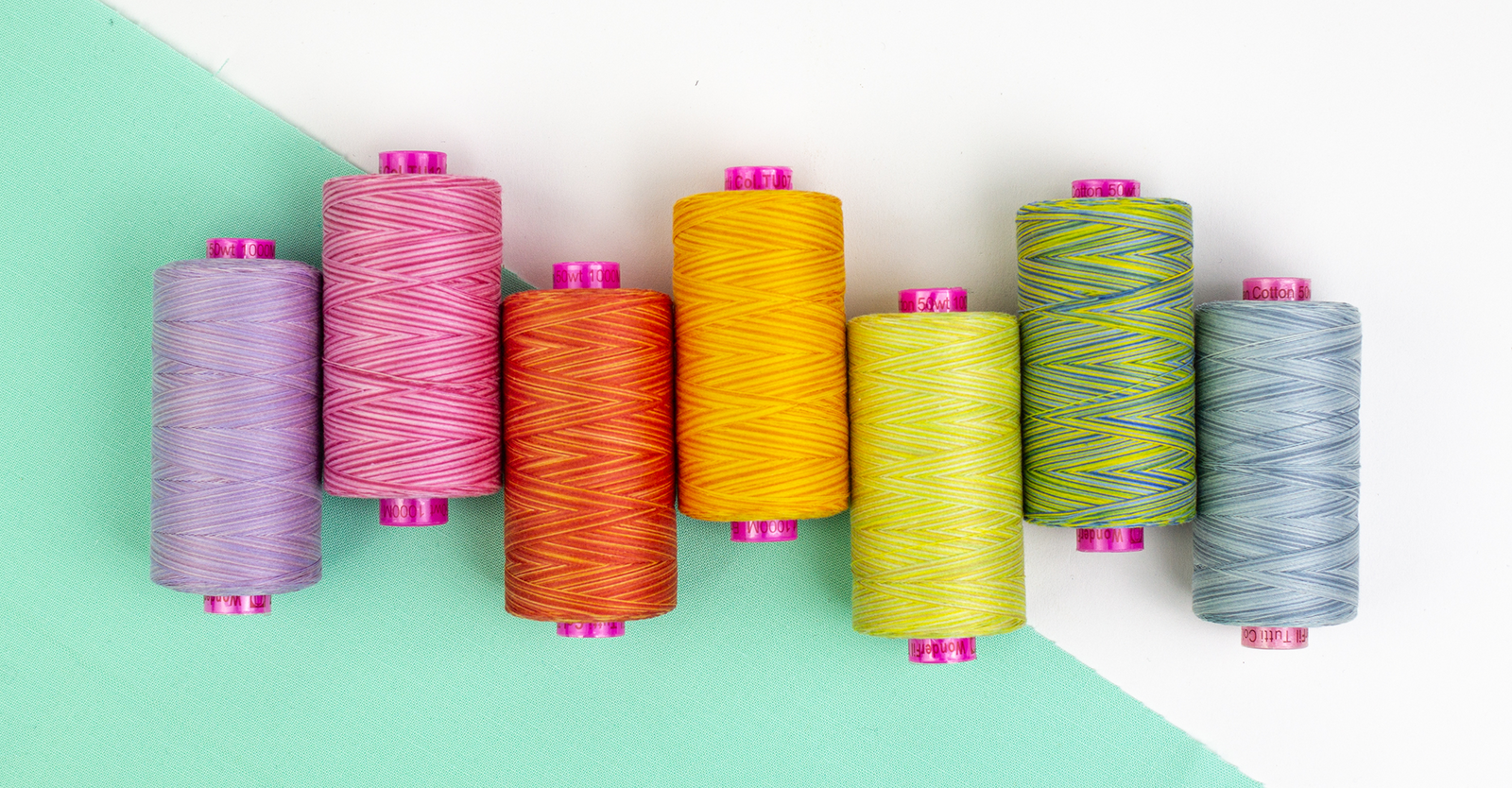 Seven spools of thread arranged vertically in a stack, each spool showing a different bright color including blue-gray, green-yellow, lime-yellow, golden yellow, orange-red, pink, and lavender. The spools have bright pink plastic cores.